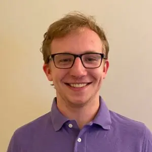 Photograph of a male with white skin. He is wearing a purple collared shirt.