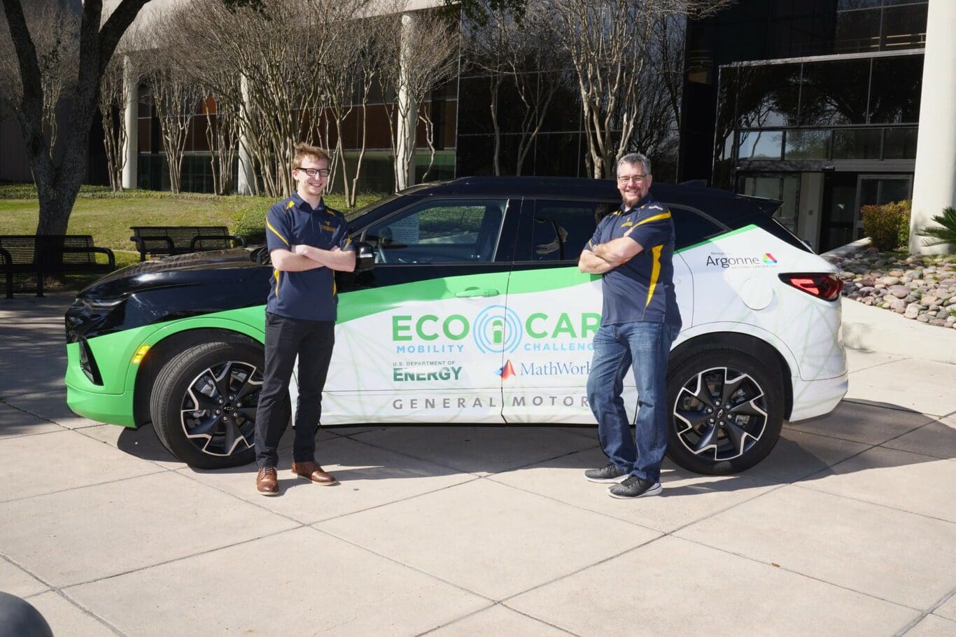 Image of two males with white skin leaning against an EcoCAR-branded vehicle. They are standing near a grassy area.